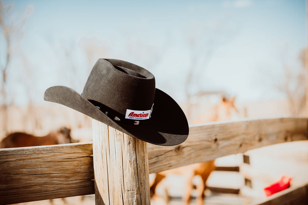 An American Brand black hat sits atop a wooden fence post on a ranch, showcasing Western Wear.